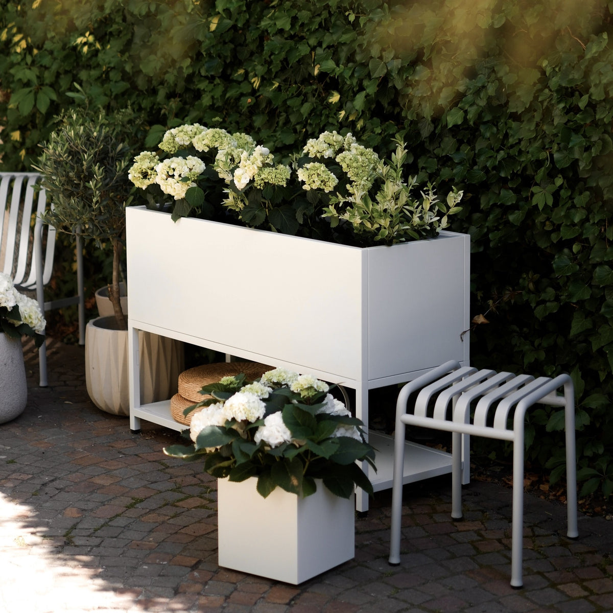 Weißes Hochbeet mit Hortensien bepflanzt und Ablagefläche, auf Terrasse zwischen Gartenstühlen, davor weißer Plantcube mit blühender Hortensie
