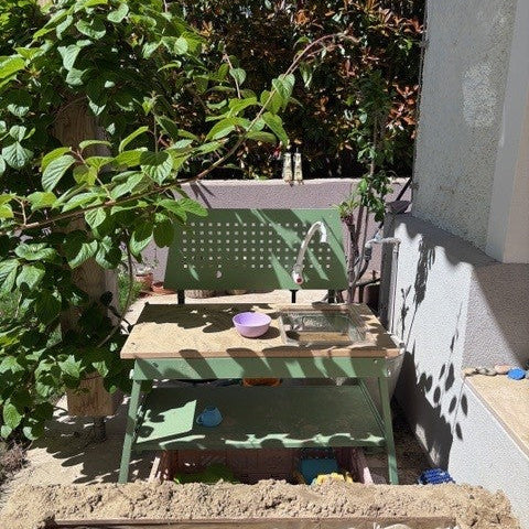 Matschküche Capri in Hellgrün hinter einem Sandkasten, links der Zweig eines Baumes