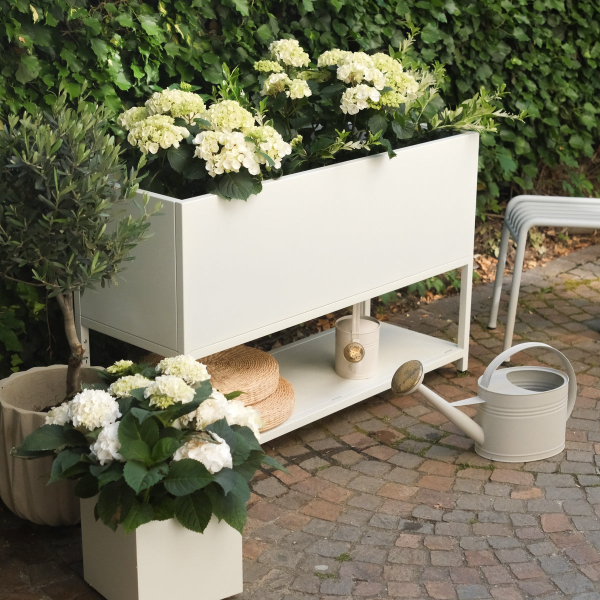 Weißes Hochbeet mit Hortensien bepflanzt darunter Ablage mit Gießkanne, davor weißer Plantcube mit blühender Hortensie auf Terrasse
